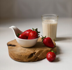 Ripe strawberries lie in a porcelain creamer and on a wooden stand , a clear glass of milk stands on a white table.