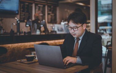 Fototapeta premium Asian businessman wearing suite and eyeglasses drink coffee , typing notebook for work at cafe restaurant. 