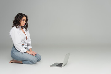 Naklejka premium Sexy brunette woman looking at laptop while sitting on grey background