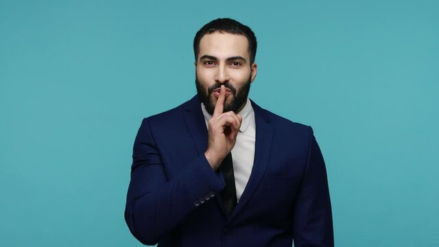 Attractive cheerful man showing shh sign holding finger near mouth, trying to keep secrets knowing interesting private information. Indoor studio shot isolated on blue background.