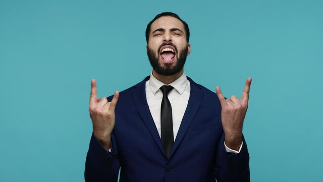 Enthusiastic crazy brunette guy in formal suit showing rock and roll sign gesture with fingers up and making grimaces, yelling happily. Indoor studio shot isolated on blue background.
