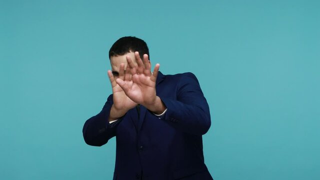 Worried scared brunette man in black suit raising his hands in defensive gesture and looking in horror at camera, walking away from frightening thing. Indoor studio shot isolated on blue background.