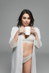 Smiling woman in silk robe and lingerie holding cup isolated on grey