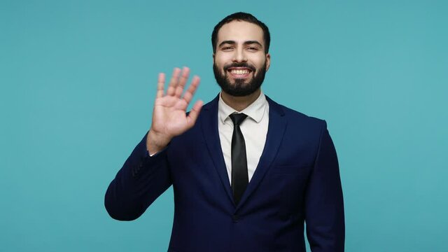 Hello! Friendly smiling happy brunette man with beard in suit saying hi, waving hand in welcome gesture, looking at camera with optimism. Indoor studio shot isolated on blue background.