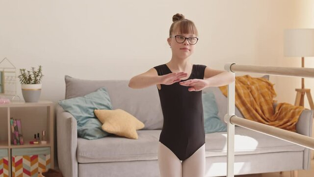 Medium slowmo of pretty elementary age girl with down syndrome in black leotard and eyeglasses doing warming-up exercise at home