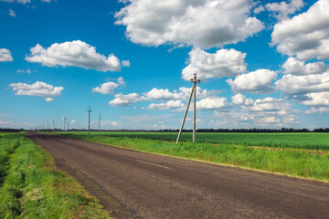 Fototapeta na wymiar Village Road with power line, green fields and blue sky with clouds