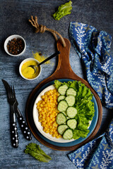 Vertical composition. Plate with salad of cucumber, corn, spices and lettuce, spoon, fork, napkin on a blue background. The concept of tasty and healthy food