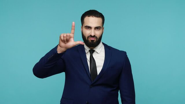 Bearded attractive businessman in suit mocking your failure, points at camera and showing L sign, blaming accusing for unsuccess, expressing disrespect. Indoor studio shot isolated on blue background.