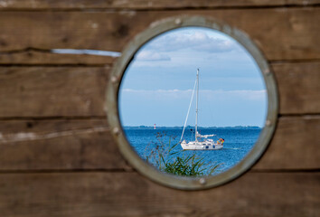 Obraz na płótnie Canvas A medium-sized sailboat in a blue sea with clouds above it. It is framed by a porthole in dark brown wood. In front there are some plants, at the horizon a lighthouse can be seen.