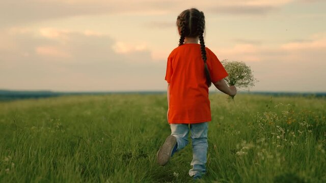 Happy child girl runs along road in green grass with bouquet of flowers for mom. Joyful, little girl dreams in nature. Childrens fantasies. Child running through field of flowers. Happy family, sunset
