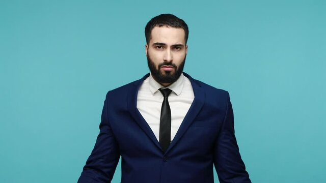 Are you crazy? Displeased brunette man with beard in black suit making stupid gesture with finger near head and pointing to camera, taunting you. Indoor studio shot isolated on blue background.