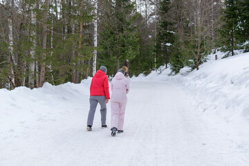 Fototapeta na wymiar Two young women go talking on a winter road with an upward slope. Coniferous green forest, white snow. View from back. Winter holiday concept, walk in park, hiking.