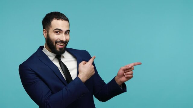 Look here! Joyful brunette bearded businessman in suit pointing copy space and nodding approvingly, showing advertising area for promotional text. Indoor studio shot isolated on blue background.