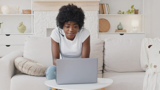 Happy African American young woman wear headset communicating by conference call speak looking at laptop at home, video chat by webcam distance language course class with computer education concept