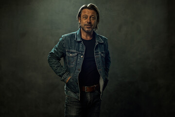Blond man in jeans with a brown leather belt and a denim jacket in front of a dark grey wall.