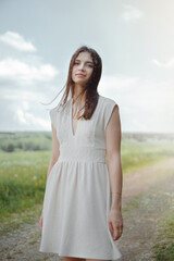 Fototapeta na wymiar Portrait of a young Caucasian brunette girl in a white dress, standing against the background of a field on a sunny summer day.