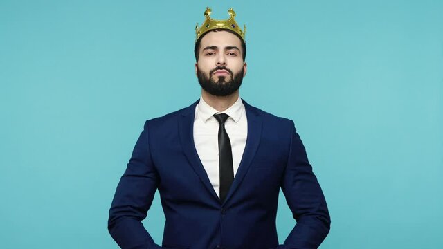 I am king! Selfish young bearded man in black formal suit wearing golden crown, symbol of power, looking with proud look, expressing confidence. Indoor studio shot isolated on blue background.