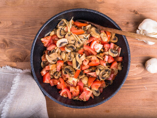 Champignon mushrooms cut into slices with onions and tomatoes are fried in a pan. Frying pan with cooking chopped mushrooms, onions and tomatoes on the table, top view, close-up