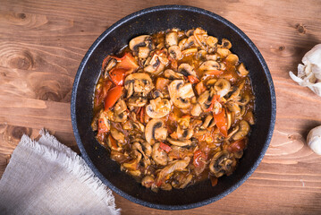 Champignon mushrooms cut into slices with onions and tomatoes are fried in a pan. Frying pan with cooking chopped mushrooms, onions and tomatoes on the table, top view., close-up