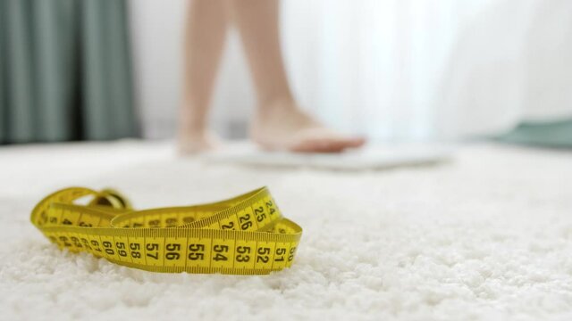 Measuring tape lying on floor next to woman standing on scales in bedroom. Concept of dieting, loosing weight and healthy lifestyle.
