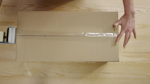 Young female online store owner, warehouse worker, merchant seller seals with tape package, preparing delivery parcel cardboard box. Hands close up. Post service. Slow motion ready 59.94fps.