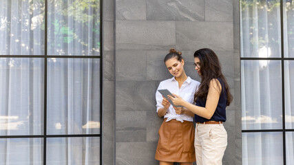 Two young attractive girls enjoy using smart phone or tablet for social connection at city sidewalk. Girls have a conversation while using tablet or mobile phone to support information © Ron