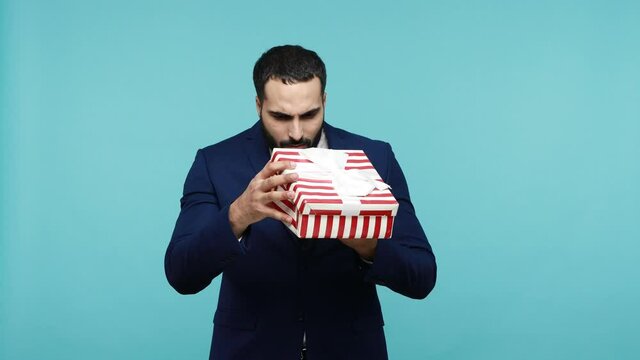 Attractive bearded businessman unpacking present box with exited facial expression, opens gift and has pleasant surprise, being satisfied. Indoor studio shot isolated on blue background.