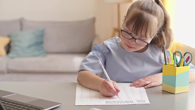 PAN waist-up shot of smart 11-year-old girl with down syndrome looking at laptop screen and doing sums on paper sitting by desk in cozy living room