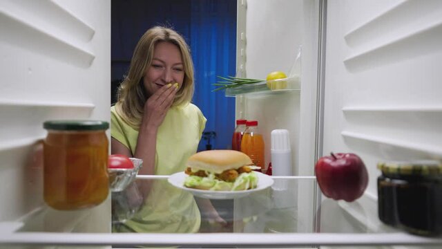 Sleepy young woman opens the refrigerator door at night, looks around with caution and is happy picks up a burger. View from inside the kitchen fridge. Close up. Slow motion ready 59.94fps.