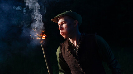 A guy dressed as Peter Pan stands with a burning torch against the night sky and dark forest