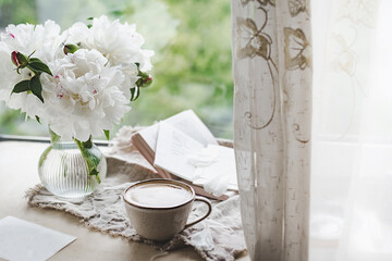 stylish ceramic cup with black coffee and foam at the window with curtains, stylish glass vase with white peonies and books. Atmospheric image of drink. cozy cafe, selective focus