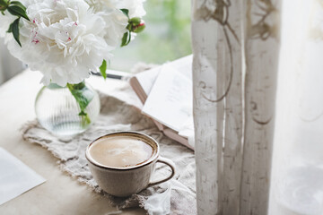 stylish ceramic cup with black coffee and foam at the window with curtains, stylish glass vase with white peonies and books. Atmospheric image of drink. cozy cafe, selective focus