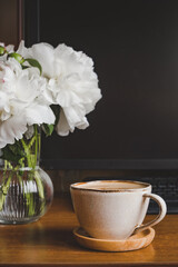 cozy home workplace. stylish cup of coffee, small glass vase with white peonies near the computer monitor. create comfort. pretty workscape. selective focus