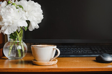 cozy home workplace. stylish cup of coffee, small glass vase with white peonies near the computer monitor. create comfort. beauty at work. selective focus