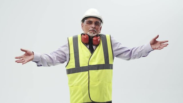 Adult man engineer in confusion spreads hands to the sides, standing in a vest and helmet with headphones on a white studio background, looking into the camera. Concept of ignorance and unknown.