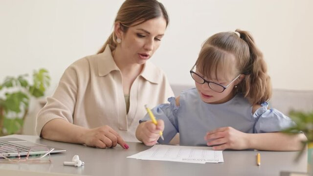 Medium slowmo of caring mother or Math teacher teaching little girl with down syndrome how to do sums sitting by desk together Girl counting on fingers and writing down right answer on piece of paper