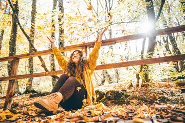 Positive curly haired woman in yellow sweater throwing dry leaves in picturesque autumn forest with colorful trees on sunny day.