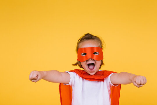 Little blonde girl dressed as a superheroine superhero with cape and red mask, screaming with open arms in flying position, on yellow background