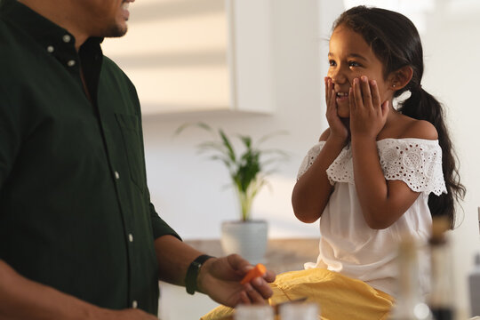 Happy hispanic daughter sitting on counter making face to father preparing vegetables in kitchen