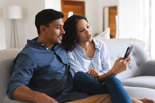 Happy hispanic couple sitting on couch in living room using smartphone