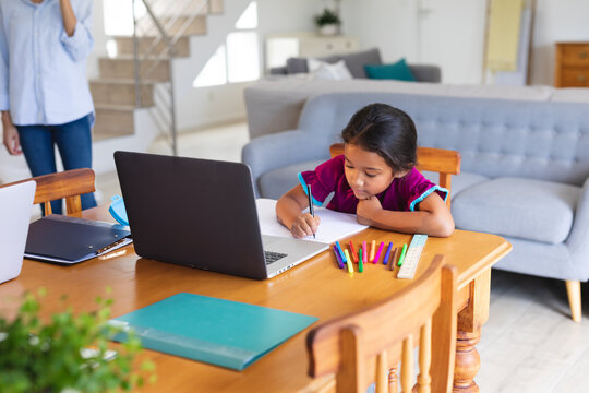 Happy hispanic girl sitting at kitchen table doing school work using laptop