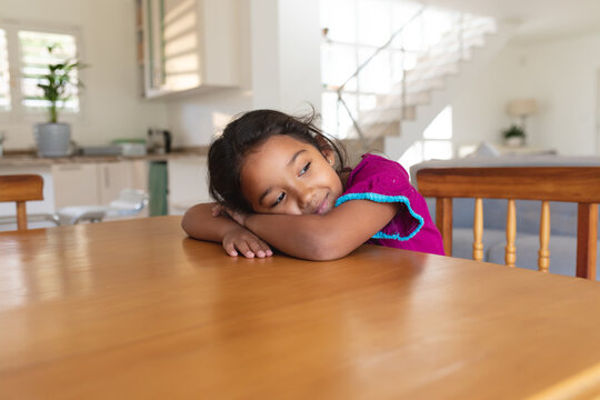 Smiling hispanic girl sitting at table in kitchen resting head on ams, looking away