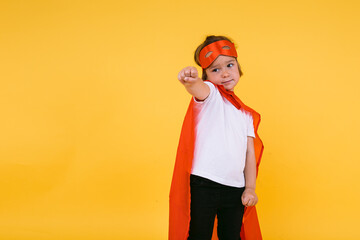 Fototapeta na wymiar Little blonde girl dressed as a superheroine superhero with a cape and red mask, raising her arm in a flying position, on yellow background