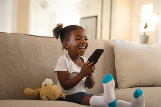 Laughing african american girl sitting on couch with teddy bear, holding remote control, watching tv