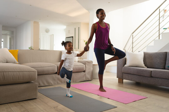 Smiling african american mother and daughter practicing yoga holding hands and standing on one leg