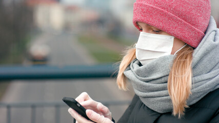 COVID-19 Coronavirus Pandemic Outbreak. Woman in Surgical Face Mask and Medical Gloves Uses Mobile App on Smartphone in Busy City Street . Close-up Picture