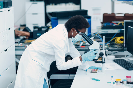 Scientist African American Black Woman In Face Mask And Gloves Working In Laboratory With Electronic Tech Instruments And Microscope.