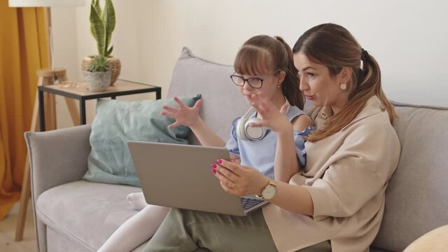 Slowmo of cheerful young mother and her elementary-age daughter with down syndrome video conferencing online on laptop while sitting on couch in bright modern living room