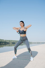 Fototapeta premium Complacent young woman exercising with resistance band on a riverside