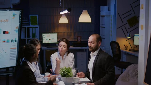Businesswoman leder sitting at desk analyzing marketing presentation discussing financial graps paperwork. Diverse multi-ethnic teamwork brainstorming project ideas working in meeting room late night